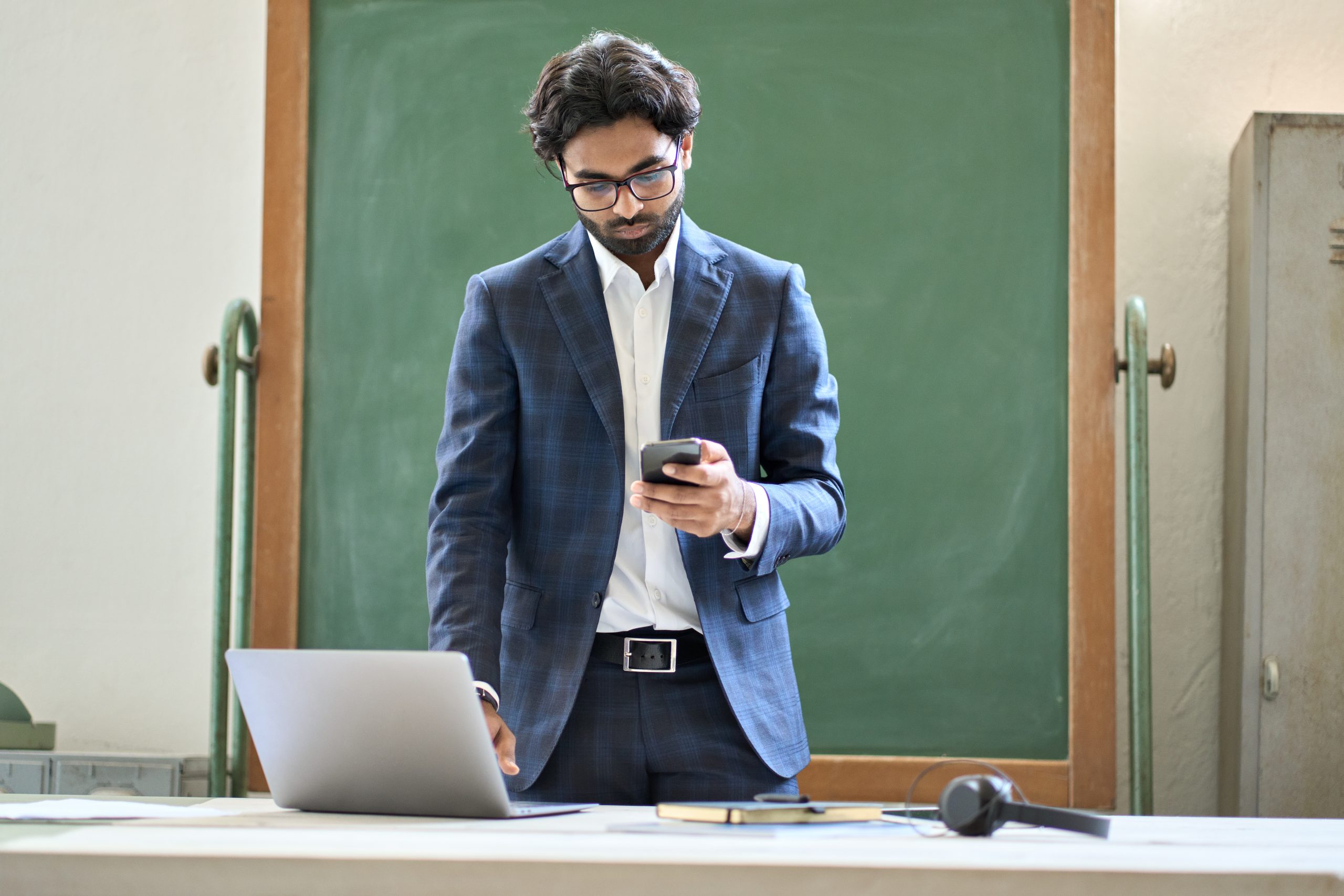 Young indian business man using phone working on laptop in office.