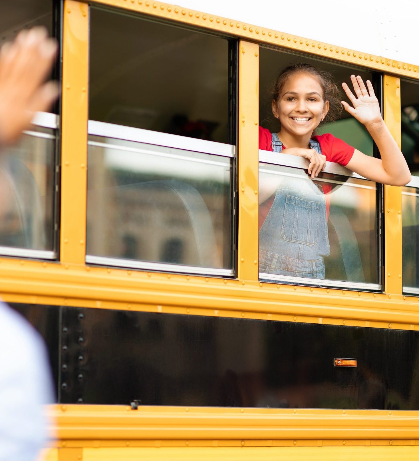 Young mom waving goodbye to happy daughter sitting in yellow school bus